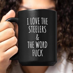 May include: A black ceramic mug featuring white text that reads I LOVE THE STEELERS & THE WORD FUCK. The mug has a standard handle and a cylindrical shape, providing a clear view of the printed message against the dark matte surface of the cup.
