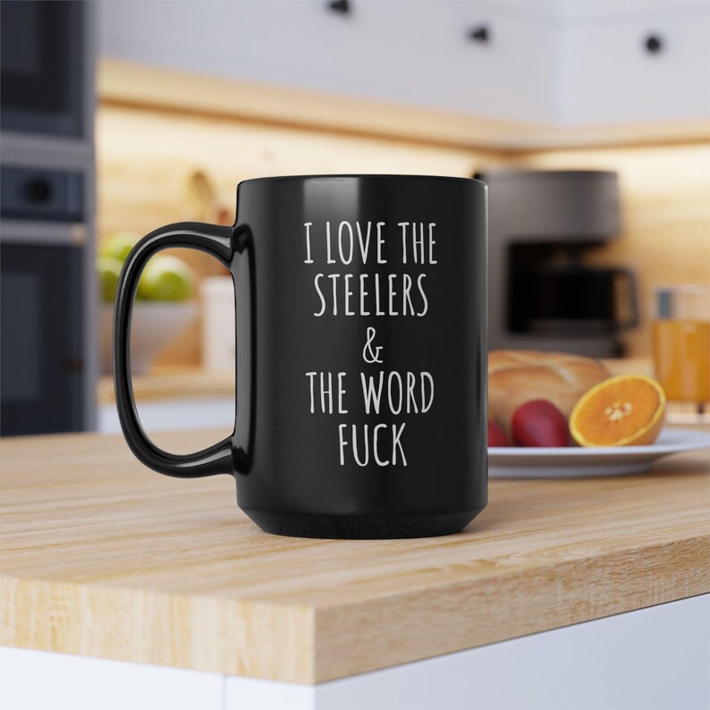 May include: A black ceramic mug with a handle on the left side. The mug features white text printed on the front that reads: I LOVE THE STEELERS & THE WORD FUCK. The mug has a smooth, matte finish and a standard cylindrical shape for holding beverages.