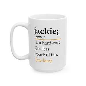 May include: A white ceramic mug featuring black text that reads: jackie; noun 1. a hard-core Steelers football fan. The text includes a gold horizontal line and the phonetic pronunciation (stē-lərz) in gold text at the bottom of the definition.