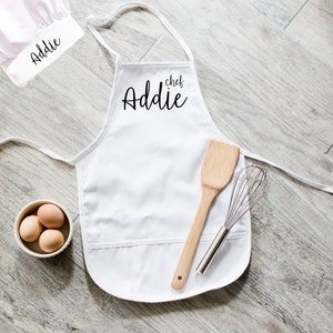 May include: White chef's hat and apron with the name "Addie chef" embroidered on the apron. The apron has a pocket and is shown with a wooden spatula and a whisk.