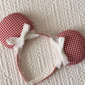 May include: A red and white gingham ear headband with white lace accents. The ears are round and padded, attached to a red and white gingham headband. A white lace bow is centered on the headband.