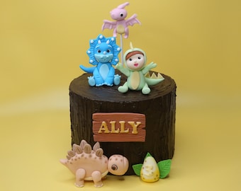 May include: A chocolate cake decorated with a wooden sign that says "ALLY" and fondant dinosaurs. The dinosaurs are a blue triceratops, a pink pterodactyl, a green dinosaur wearing a costume, and a brown stegosaurus.
