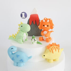May include: A cake decorated with fondant dinosaurs, a volcano, and a number one. The dinosaurs are green, orange, blue, and yellow. The volcano is gray with red lava.