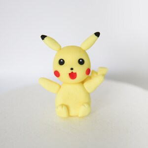 Puede incluir: Un personaje de Pikachu amarillo hecho de fondant, con orejas y ojos negros, y mejillas rojas. El personaje est&aacute; sentado con los brazos extendidos.