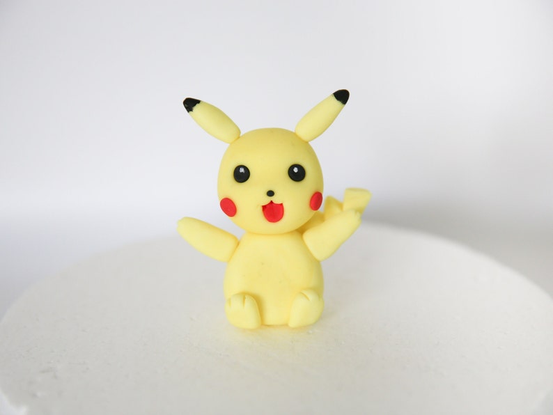 Puede incluir: Un personaje de Pikachu amarillo hecho de fondant, con orejas y ojos negros, y mejillas rojas. El personaje est&aacute; sentado con los brazos extendidos.