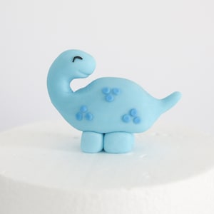 May include: A light blue fondant dinosaur cake topper with small blue dots. The dinosaur has a smiling face and is standing on two legs.