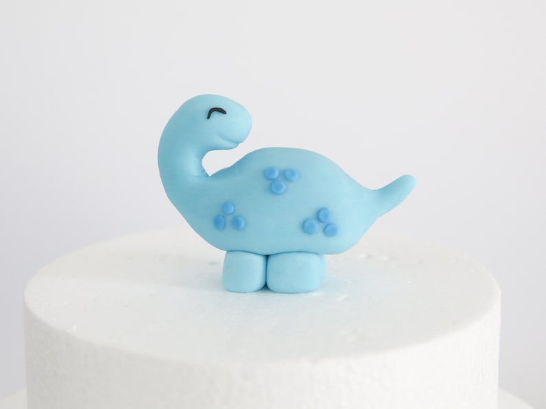 May include: A light blue fondant dinosaur cake topper with small blue dots. The dinosaur has a smiling face and is standing on two legs.