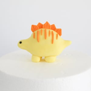 May include: A yellow fondant dinosaur cake topper with an orange spiky back and small black eyes.