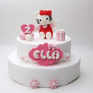 May include: Two-tiered white cake decorated with a pink and white Hello Kitty figurine, a pink heart with the number 2, pink and white flowers, and a pink sign that says "Ella".