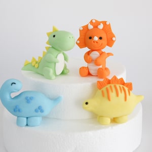 May include: Four colorful dinosaur cake toppers made of fondant. The dinosaurs are green, orange, blue, and yellow. They are all in a playful, cartoon style.