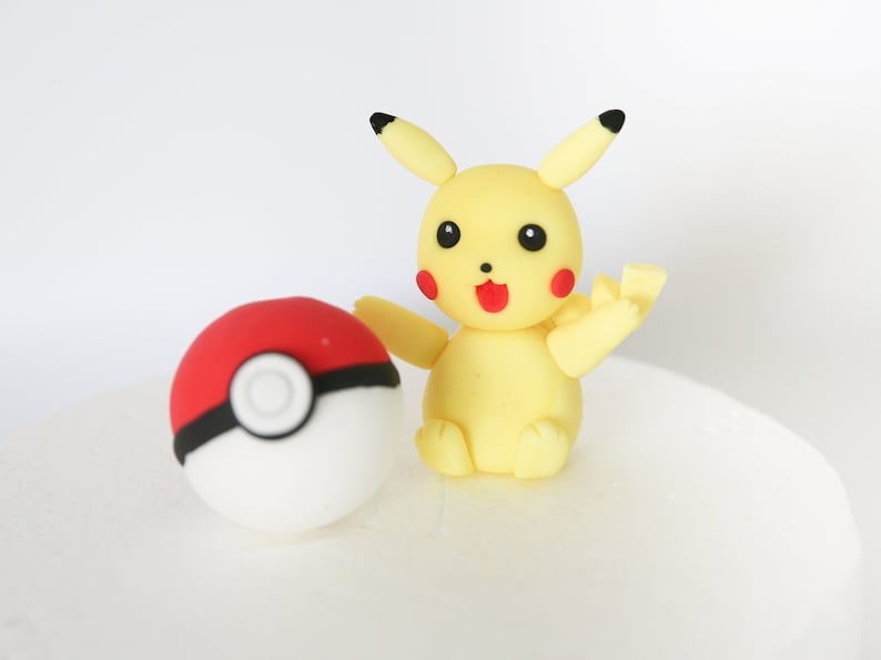 Puede incluir: Una figura de Pikachu amarilla con mejillas rojas y nariz negra, sosteniendo una Pok&eacute; Ball roja y blanca con una banda negra.