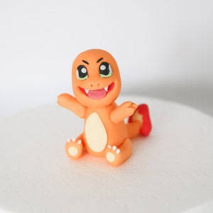 Puede incluir: Topper de pastel de fondant naranja y amarillo de un personaje de dibujos animados con una amplia sonrisa y ojos grandes. El personaje tiene una cola roja y est&aacute; sentado sobre sus ancas.