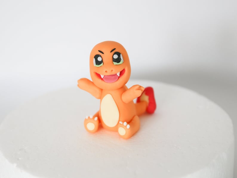 Puede incluir: Topper de pastel de fondant naranja y amarillo de un personaje de dibujos animados con una amplia sonrisa y ojos grandes. El personaje tiene una cola roja y est&aacute; sentado sobre sus ancas.