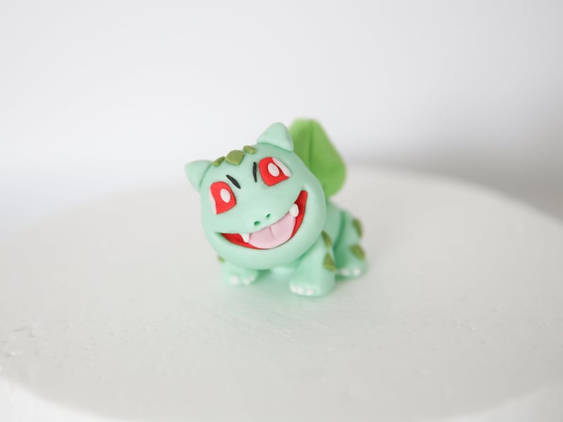 Puede incluir: Una figurita de fondant verde y roja de un personaje de dibujos animados con una amplia sonrisa y ojos grandes. La figurita es un personaje popular de una serie de anime y videojuegos japoneses.