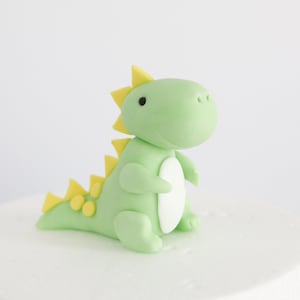 May include: A green and yellow fondant dinosaur cake topper. The dinosaur has a friendly face and a spiky back.