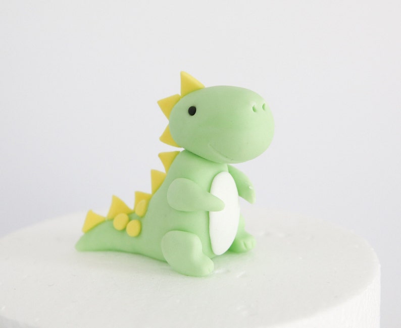 May include: A green and yellow fondant dinosaur cake topper. The dinosaur has a friendly face and a spiky back.