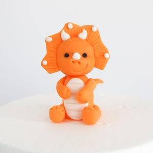 May include: Orange and white fondant triceratops dinosaur cake topper. The dinosaur has a friendly face and is sitting on its hind legs.