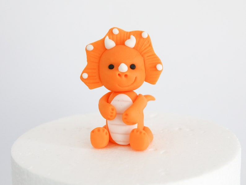 May include: Orange and white fondant triceratops dinosaur cake topper. The dinosaur has a friendly face and is sitting on its hind legs.