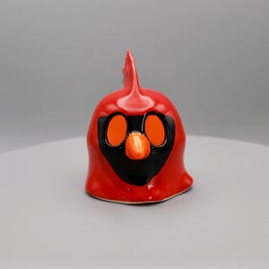 May include: A ceramic cardinal figurine in bright red with black and orange details. The bird has a prominent crest, large orange eyes, and a small orange beak. The figurine is a decorative item.