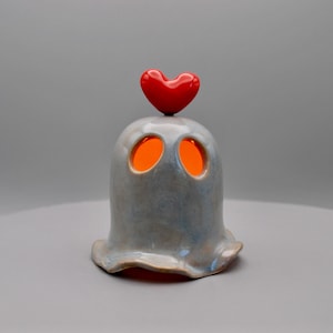 May include: A ceramic ghost-shaped candle holder with a red heart on top. The ghost is light blue with orange glowing eyes. The base has a ruffled edge. A unique decorative item.