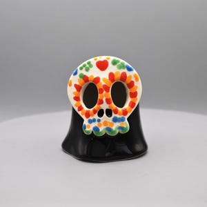 May include: A Day of the Dead-themed ceramic skull figurine. The skull is white with colorful floral and heart designs. The base is black and has a glossy finish. The figurine is a decorative item.