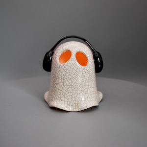 May include: A ceramic ghost figure with a cracked white finish and orange eye-holes. The ghost is wearing black headphones. The figure is a Halloween or spooky-themed decorative item.
