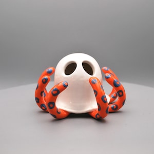 Pode incluir: Um objeto decorativo de cerâmica branca em forma de polvo, com tentáculos laranja com bolinhas azuis. O polvo tem dois buracos para os olhos. Ideal para decoração de interiores.