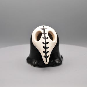 May include: A ceramic figurine of a plague doctor mask. The mask is off-white with a black vertical line design. The base is black and the figurine has a glossy finish. The figurine is on a gray surface.