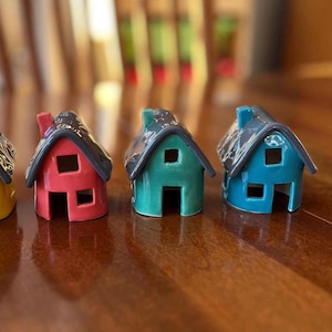 Könnte beinhalten: Nahaufnahme von drei keramischen Miniaturhäusern in Rot, Türkis und Blau. Jedes Haus hat ein dunkelgraues Dach und eine rechteckige Fenster- und Türöffnung. Die Häuser sind auf einer Holzoberfläche angeordnet.
