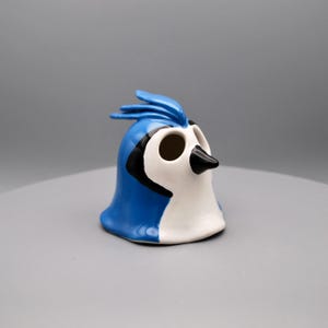 Puede incluir: Figura de cerámica de arrendajo azul con vientre blanco y detalles negros. El ave tiene una cresta distintiva y un pico negro. La figura es un elemento decorativo.