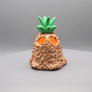 May include: A whimsical, handcrafted pineapple-shaped decorative object. The body is textured with a brown, speckled finish, and features two orange, glowing eye-like openings. The top has a vibrant green, leafy crown. This unique piece adds a touch of fun.