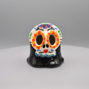 May include: A ceramic skull figurine with a black base and a white skull face. The skull face is decorated with orange, blue, green, and red floral patterns. The figurine is a decorative item.