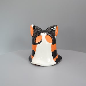 May include: A ceramic cat-shaped ornament in black, orange, and white. The figurine has a rounded base and pointed ears. The cat's face features orange eye markings and a white muzzle. A decorative item.