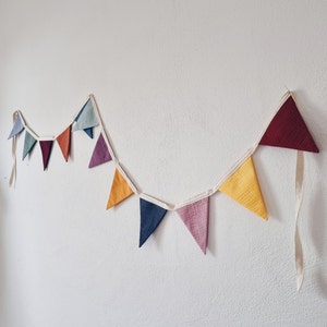 May include: A colorful fabric bunting with triangular flags in shades of red, orange, yellow, green, blue, purple, and brown. The bunting is strung on a white ribbon and is hanging on a white wall.