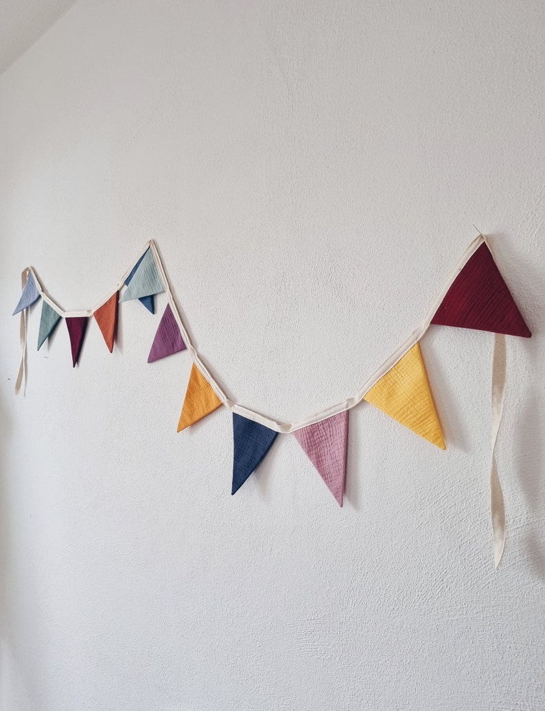 May include: A colorful fabric bunting with triangular flags in shades of red, orange, yellow, green, blue, purple, and brown. The bunting is strung on a white ribbon and is hanging on a white wall.