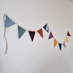 May include: A colorful fabric bunting with triangular flags in shades of blue, green, red, orange, yellow, and purple. The bunting is strung on a white cord and is hanging on a white wall.