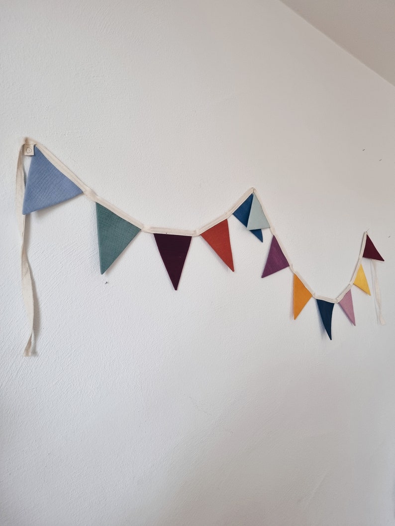 May include: A colorful fabric bunting with triangular flags in shades of blue, green, red, orange, yellow, and purple. The bunting is strung on a white cord and is hanging on a white wall.