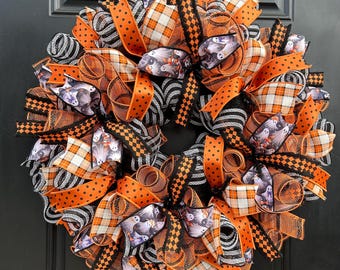 May include: A decorative Halloween wreath featuring a mix of orange, black, and white mesh ribbons. The design incorporates patterned ribbons with black polka dots, orange and white plaid, black and white stripes, and a ghost-themed print.