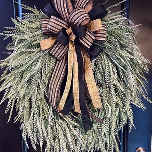 May include: A decorative wreath featuring a base of green, feathery foliage. The wreath is adorned with a large bow composed of black, gold, and striped ribbons. The wreath is hanging on a black door.