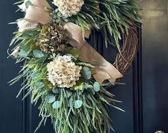 May include: A green and white wreath with a hessian bow. The wreath is made of greenery and has white hydrangeas and green flowers.