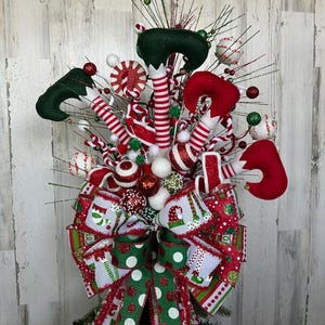 May include: A festive Christmas tree topper featuring elf legs, candy canes, and decorative balls in red, white, and green. A large bow with elf and polka dot patterns adds a whimsical touch to this holiday decoration.