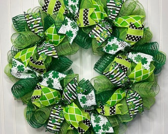 Op de afbeelding: Een feestelijke St. Patrick's Day krans met een cirkelvormig ontwerp. De krans is gemaakt van groen gaas en versierd met linten in groen, wit en zwart. De linten tonen patronen zoals stippen, strepen en klavers.