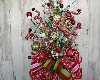 May include: A festive Christmas tree topper featuring red, green, and white ornaments, candy canes, and a large bow. The bow has red fabric with white stripes and green accents. The topper is decorated with glitter.