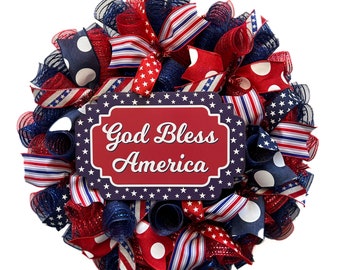 May include: A red, white, and blue patriotic wreath with a sign that reads "God Bless America". The wreath is made of mesh and ribbon and features polka dots and stars.