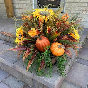 May include: A fall-themed arrangement featuring sunflowers, pumpkins, and various foliage. The arrangement is in a wooden barrel-shaped container, with orange pumpkins and yellow sunflowers as focal points. The arrangement is placed on a gray brick step.