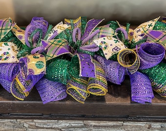 May include: A Mardi Gras themed door swag with purple, green, and gold mesh, ribbons, and patterned fabric. The swag is decorated with Mardi Gras beads and a gold star.