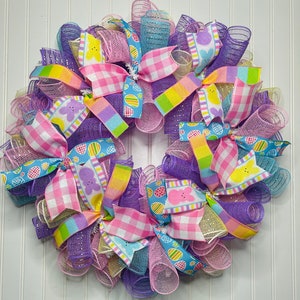 May include: A colorful Easter wreath with purple, pink, blue, yellow, and white mesh and ribbon. The ribbon features a checkered pattern and a design of pastel-colored eggs and bunnies.