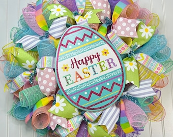 Puede incluir: Una corona de Pascua colorida con un letrero en forma de huevo turquesa que dice "Feliz Pascua" en letras rosas y blancas. La corona está hecha de malla y cinta en tonos de rosa, morado, verde, amarillo y blanco. Presenta margaritas blancas y patrones de lunares.