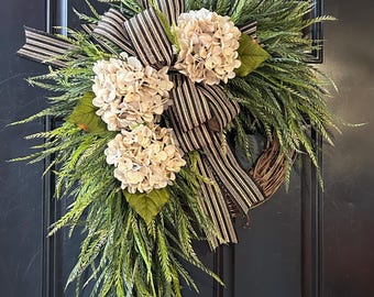 May include: A wreath with a black and white striped ribbon and white hydrangea flowers. The wreath is made of green foliage and is hanging on a black door.