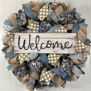 May include: A burlap wreath with blue and white checkered ribbon, brown polka dot ribbon, and black damask ribbon. A wooden sign with the word "Welcome" is in the center of the wreath.